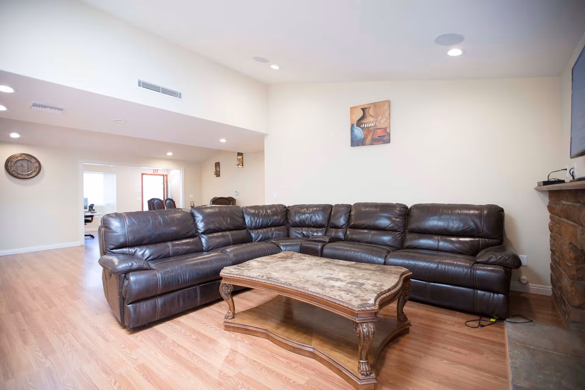 A spacious living room with a large dark brown leather sectional sofa and a wooden coffee table with a marble top. The room has light-colored walls, wood flooring, recessed ceiling lights, a wall clock, a painting, and a stone fireplace with a mounted TV above it.