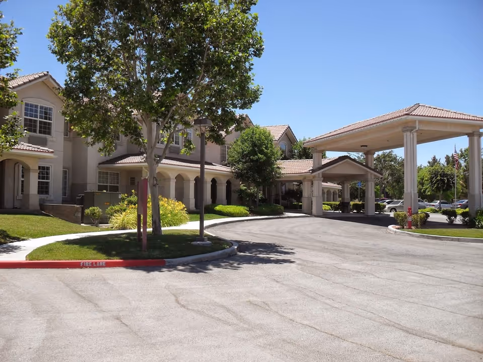 Exterior view of Bellamar Lancaster facility showing a two-story building with beige walls and tiled roof, surrounded by trees and bushes. There is a covered entrance with columns and a driveway in front, under a clear blue sky.