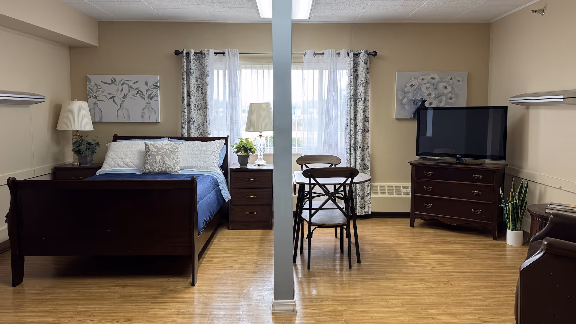 Furnished assisted-living studio room with a bed, nightstands and lamps, a small dining table, a TV on a dresser, and a window with curtains.