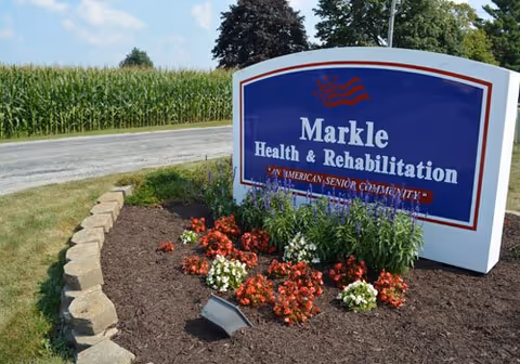 A blue sign reading "Markle Health & Rehabilitation" stands in a landscaped flower bed beside a road with a cornfield in the background.