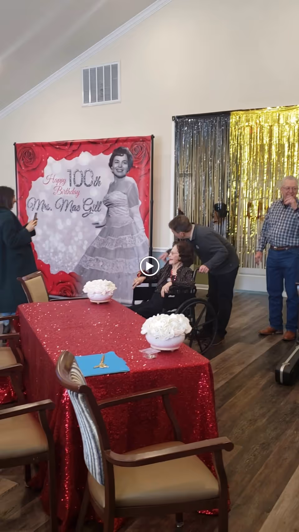 Decorated common room with a red-sequined table, floral centerpieces, and residents gathered near a 100th birthday backdrop.