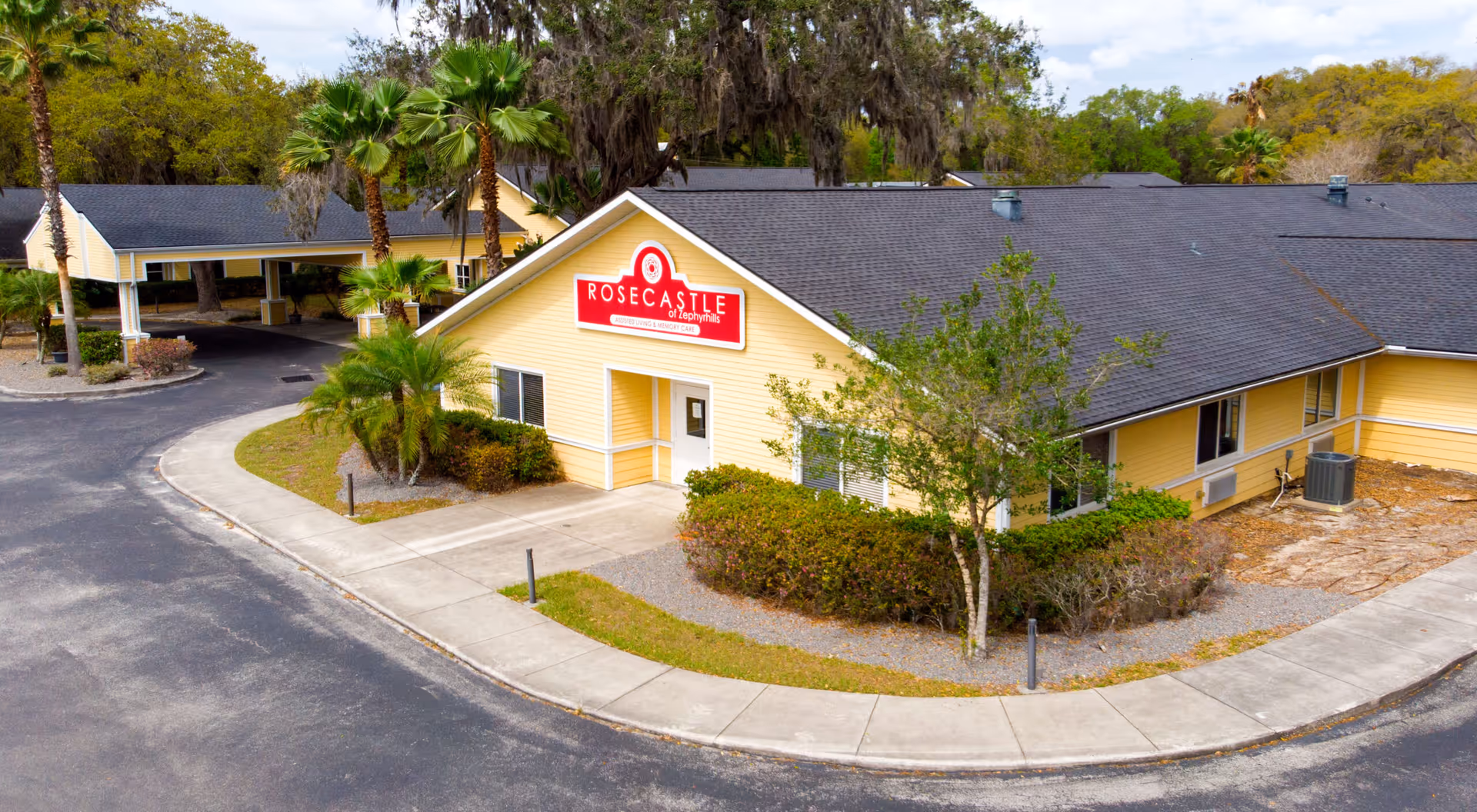Exterior view of a single-story yellow building with a dark roof, surrounded by palm trees and bushes. A red sign on the building reads 'ROSECASTLE of Zephyrhills'. The building is situated along a curved driveway with a covered entrance area visible in the background.