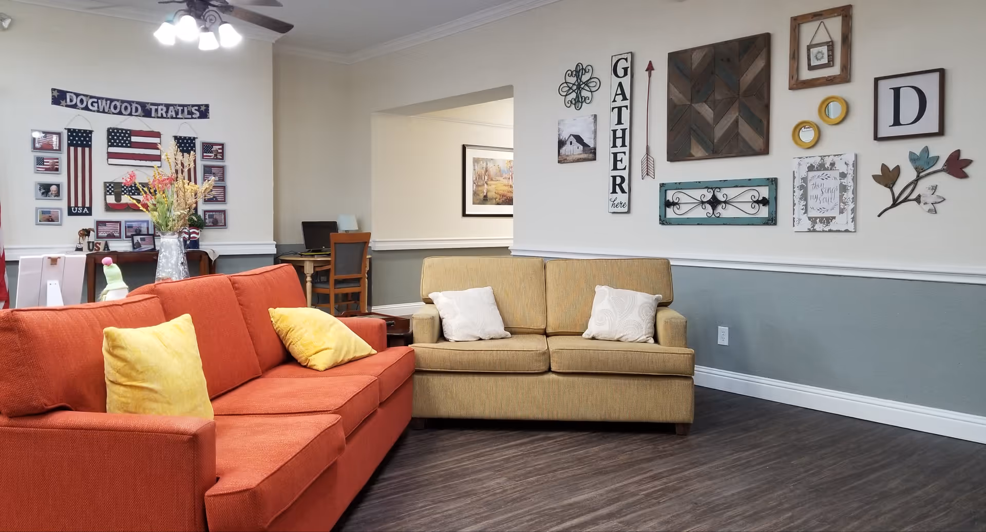 A bright assisted living common room with red and beige sofas, decorative wall art including a 'Dogwood Trails' sign, and a small desk in the background.