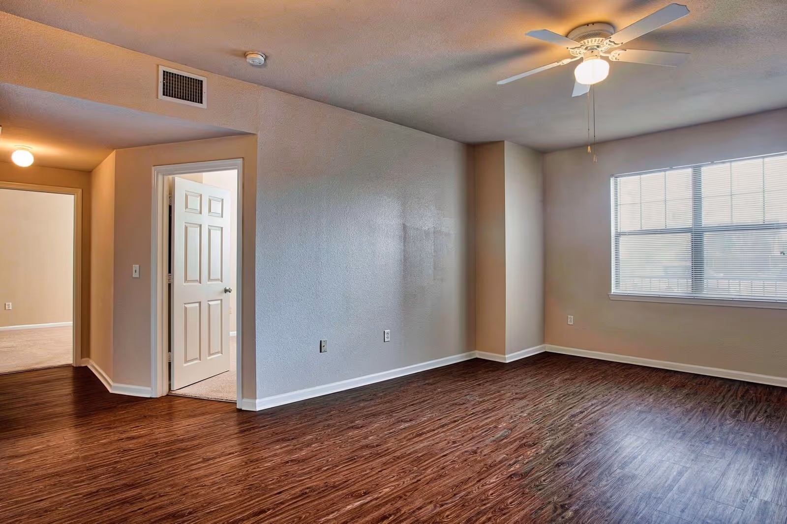 Empty room with wood flooring, beige walls, a ceiling fan with light, and a large window with blinds. There is an open door leading to a carpeted room and a hallway with a light fixture.