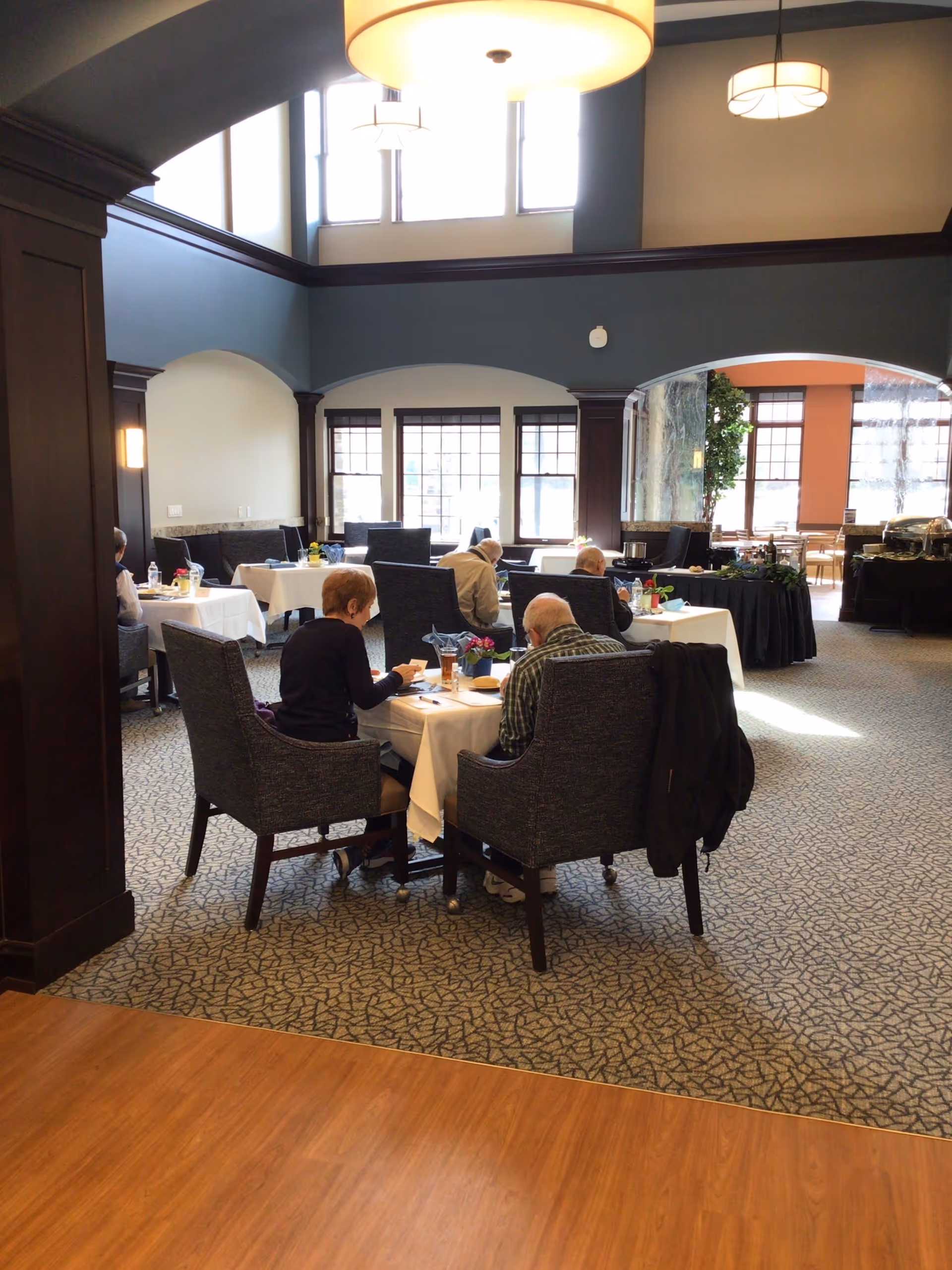 Several elderly residents seated at a table in a spacious, high-ceiling dining room with large windows and pendant lights.