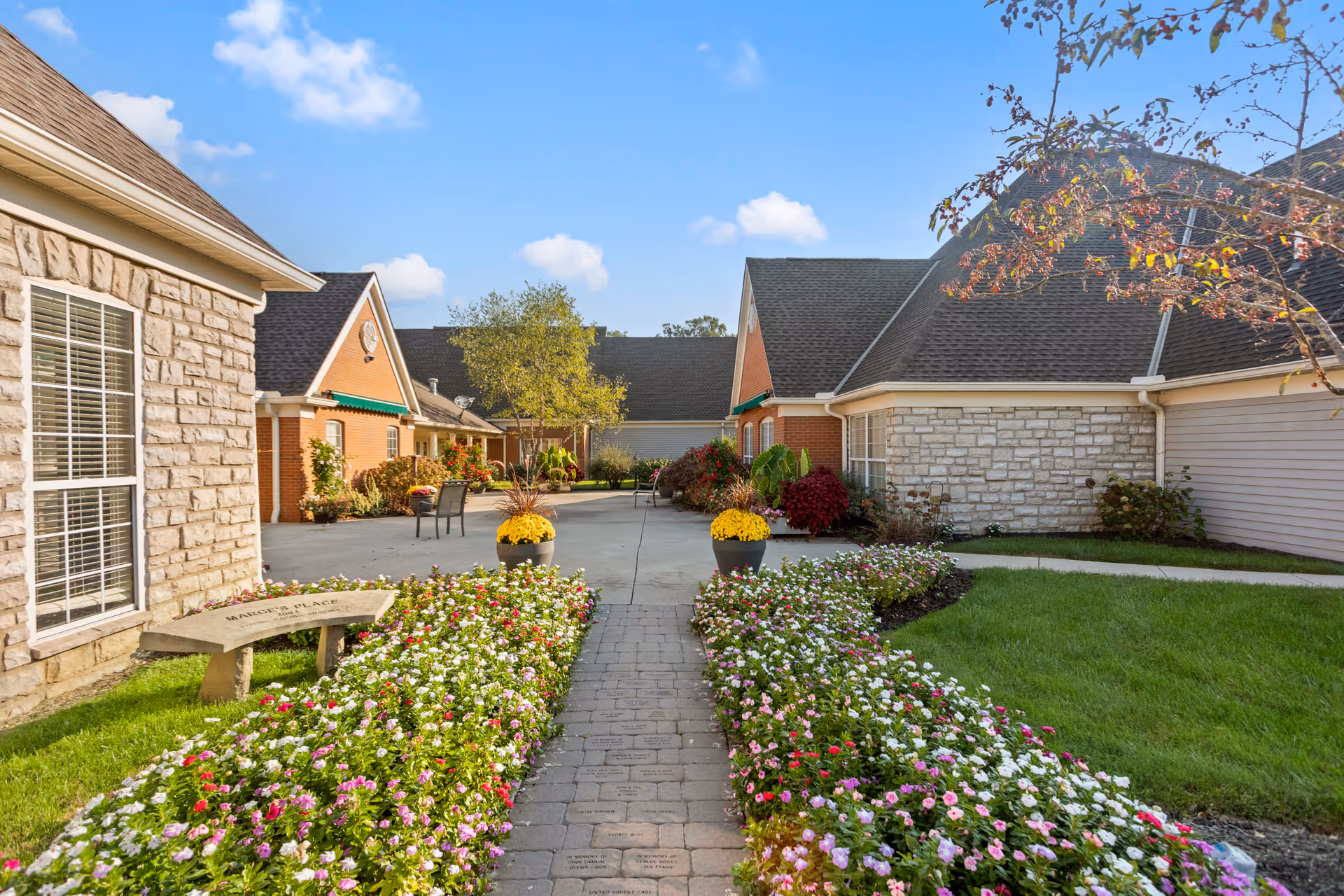 A bright outdoor courtyard area at The Gables of Marysville featuring a paved walkway lined with colorful flower beds and two large flower pots with yellow flowers. The courtyard is surrounded by buildings with brick and stone exteriors, green awnings, and a clear blue sky overhead.