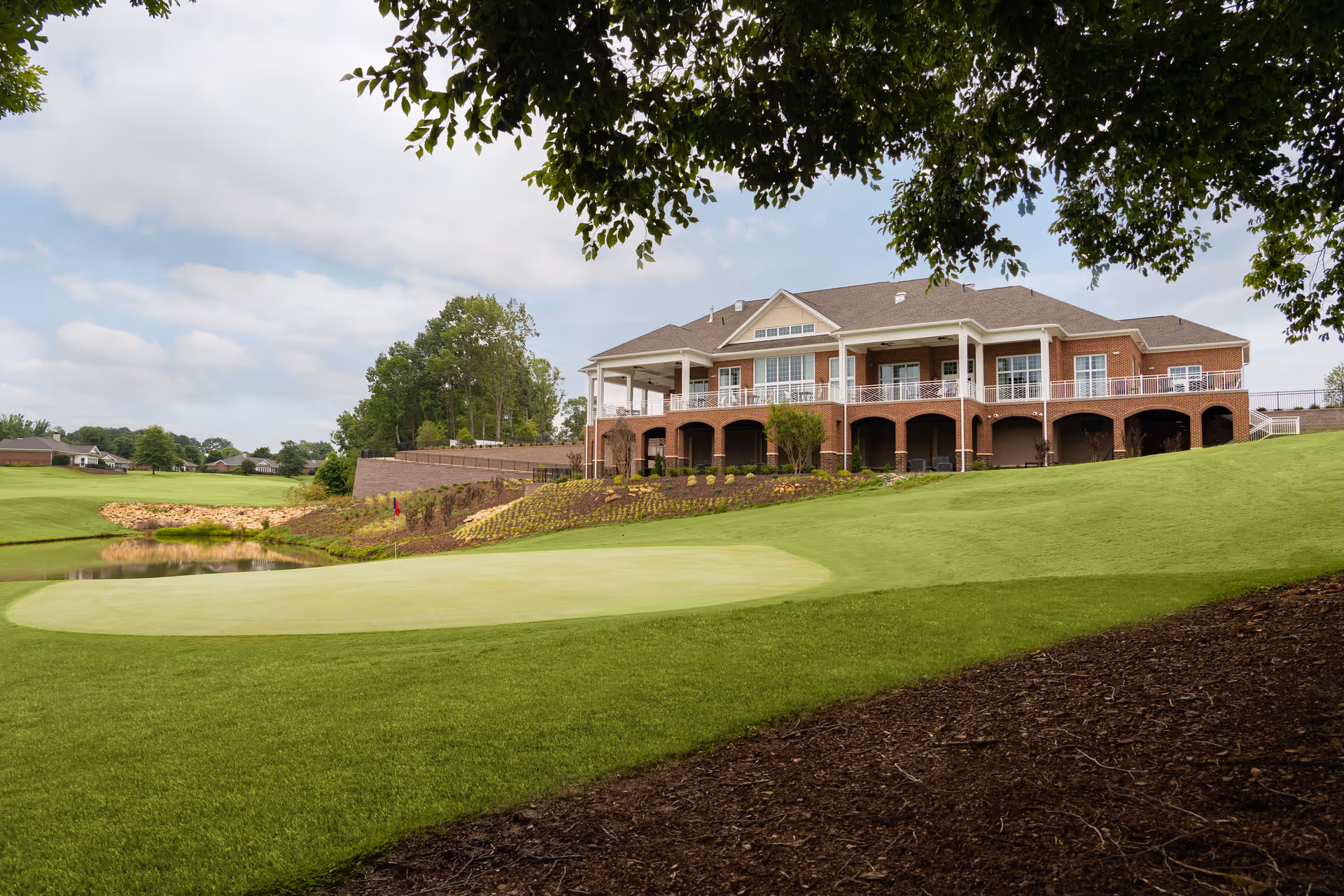 Brick senior living building with covered balconies overlooking a golf green and pond framed by tree branches.