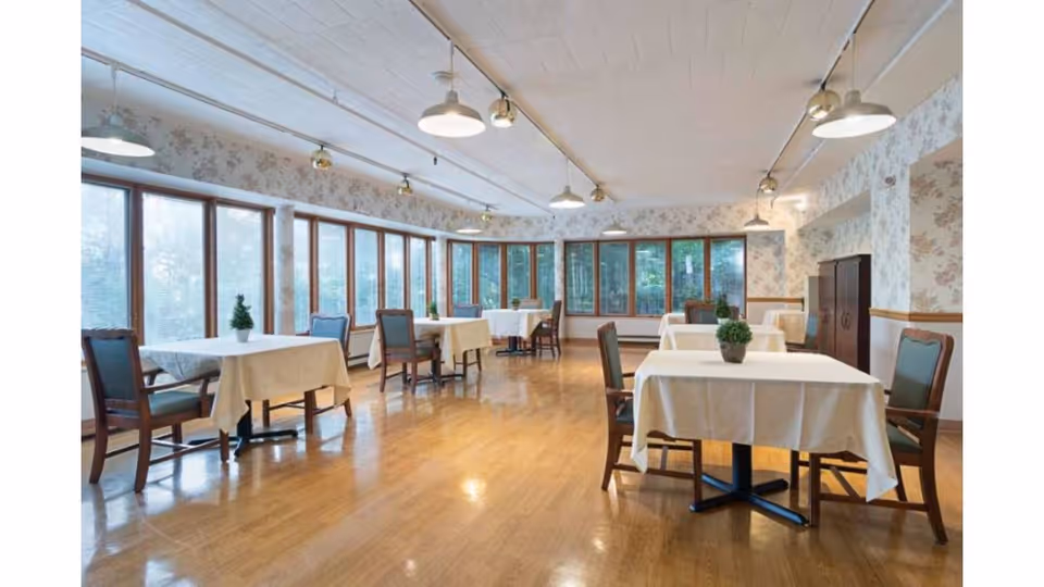 A bright dining room with large windows along two walls, letting in natural light. The room has wooden floors and floral wallpaper. Several tables are covered with white tablecloths, each accompanied by wooden chairs with green cushions. Small potted plants are placed on each table.
