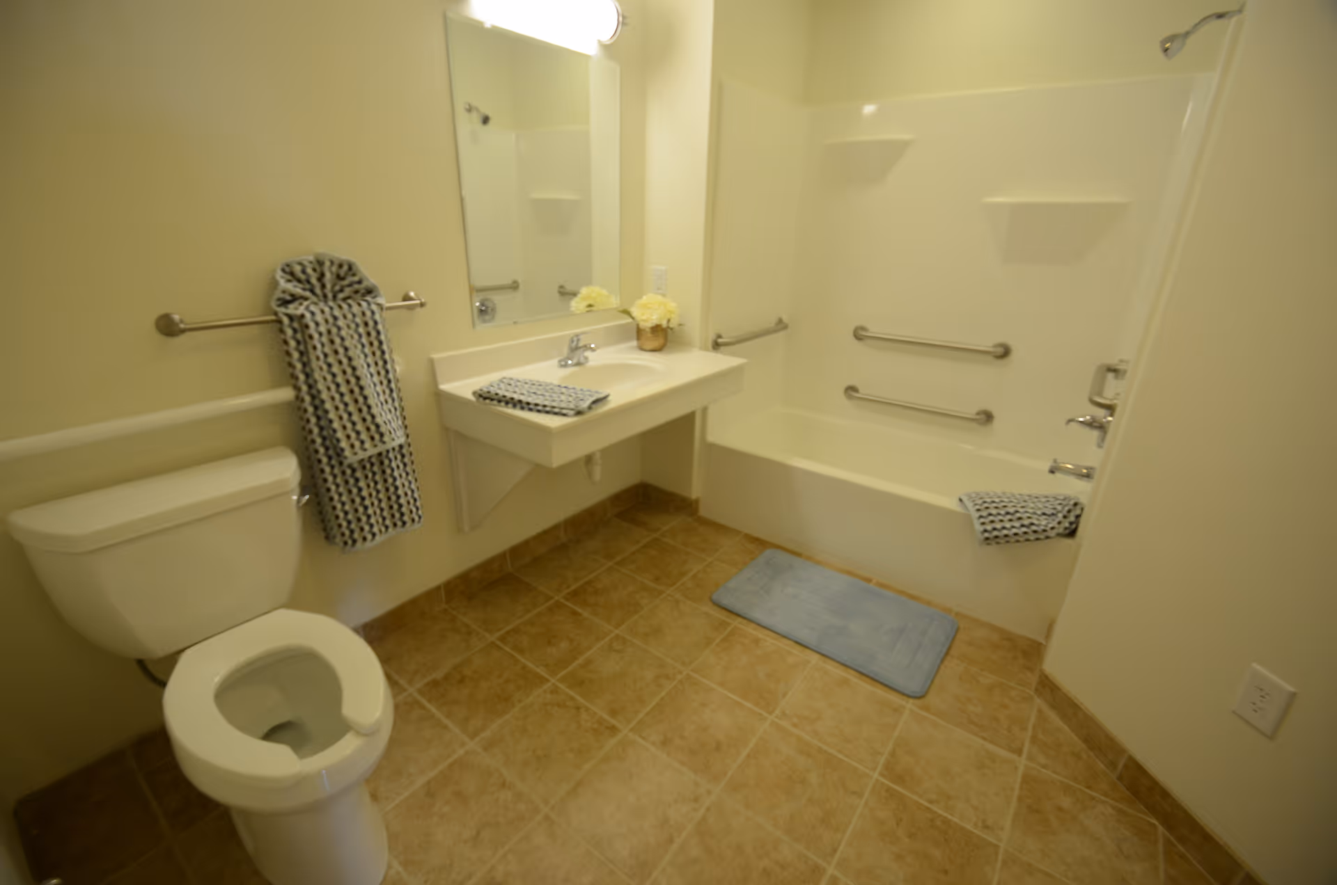 Accessible bathroom with a toilet, wall-mounted sink and mirror, bathtub with grab bars, towels, and a tiled floor.