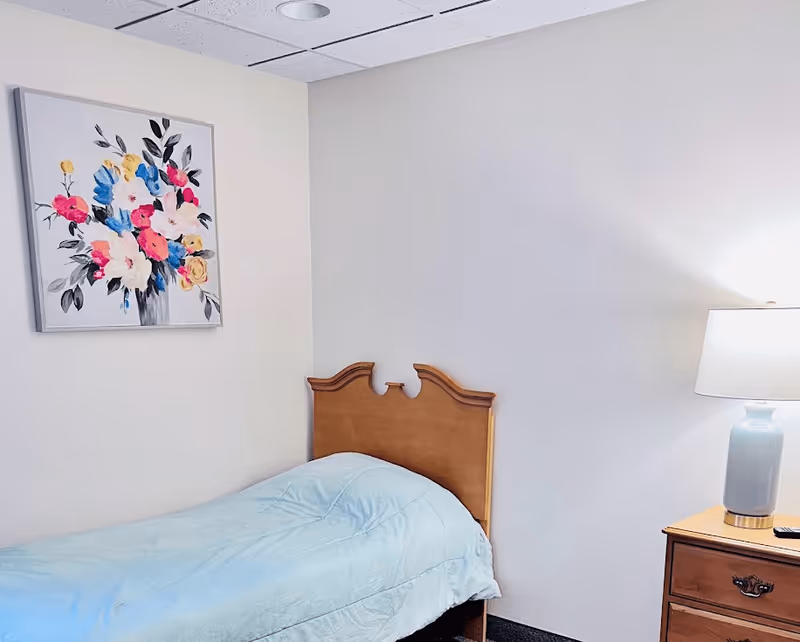 A simple bedroom with a single wooden bed with a light blue comforter, a wooden nightstand with a blue lamp, and a colorful floral painting on the wall.