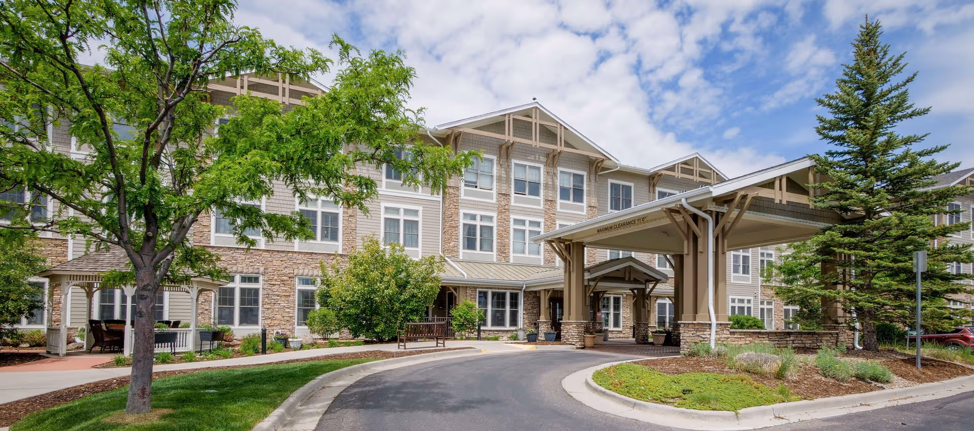 Exterior view of a senior living facility building with a covered entrance, stone and siding facade, multiple windows, landscaped greenery including trees and bushes, and a curved driveway under a partly cloudy sky.