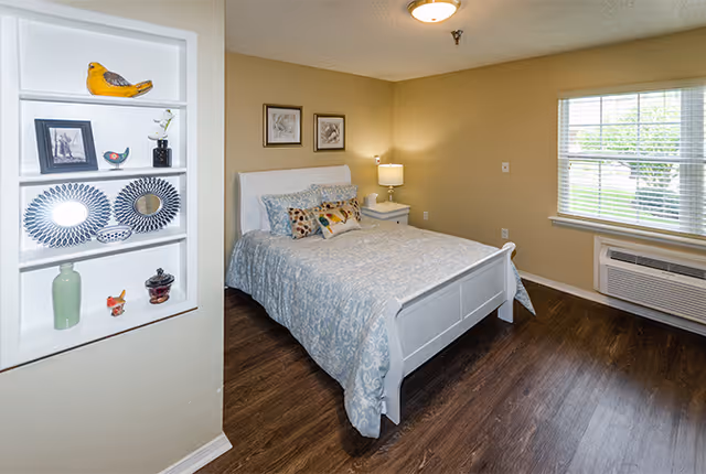 A cozy bedroom with a white wooden bed frame and a light blue patterned bedspread. The bed has several decorative pillows. Next to the bed is a white nightstand with a lamp. The room has wood flooring and beige walls. A window with blinds lets in natural light, and an air conditioning unit is installed below the window. On the left wall, there is a built-in white shelf displaying decorative items including plates, a yellow bird figurine, and small vases.
