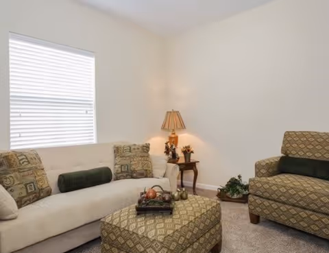 A cozy living room with a beige sofa, patterned armchair and ottoman, a side table with a lamp, and a window with blinds.