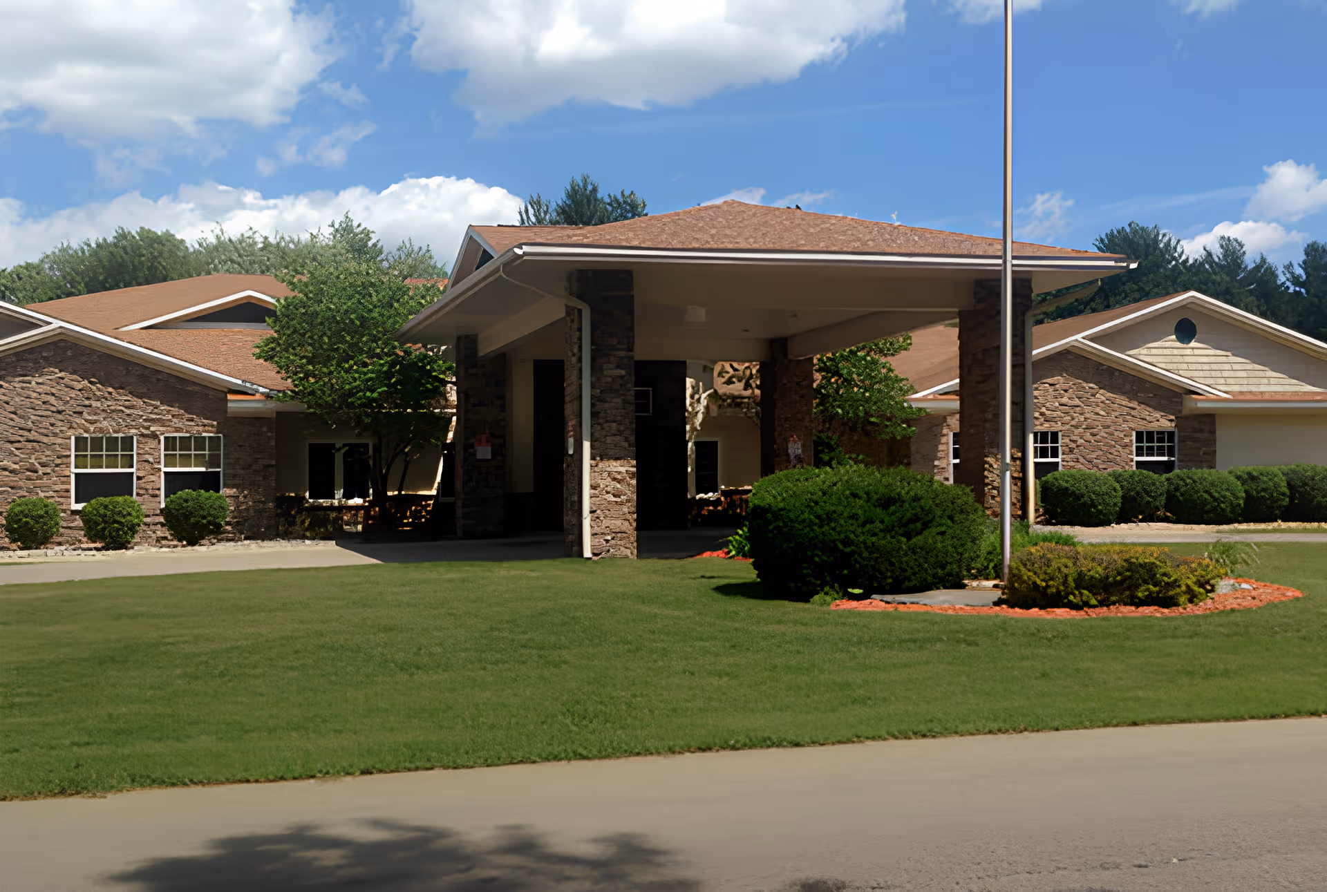 Front entrance of a senior living building with a covered porte-cochère, stone facade, manicured lawn and flagpole.