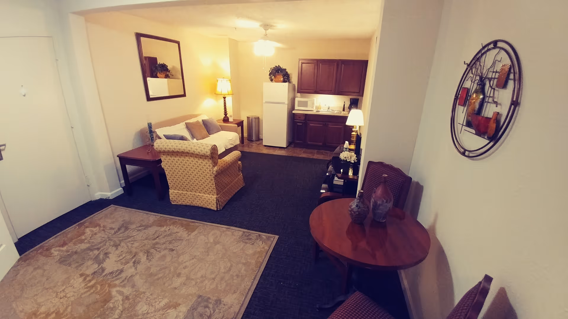 Interior view of a senior living apartment featuring a small living area with a couch, armchair, side table with a lamp, and a mirror on the wall. In the background, there is a kitchenette with a refrigerator, microwave, coffee maker, and cabinets. A round wooden table with two chairs and decorative vases is positioned near the foreground. The room is warmly lit with lamps and ceiling light.