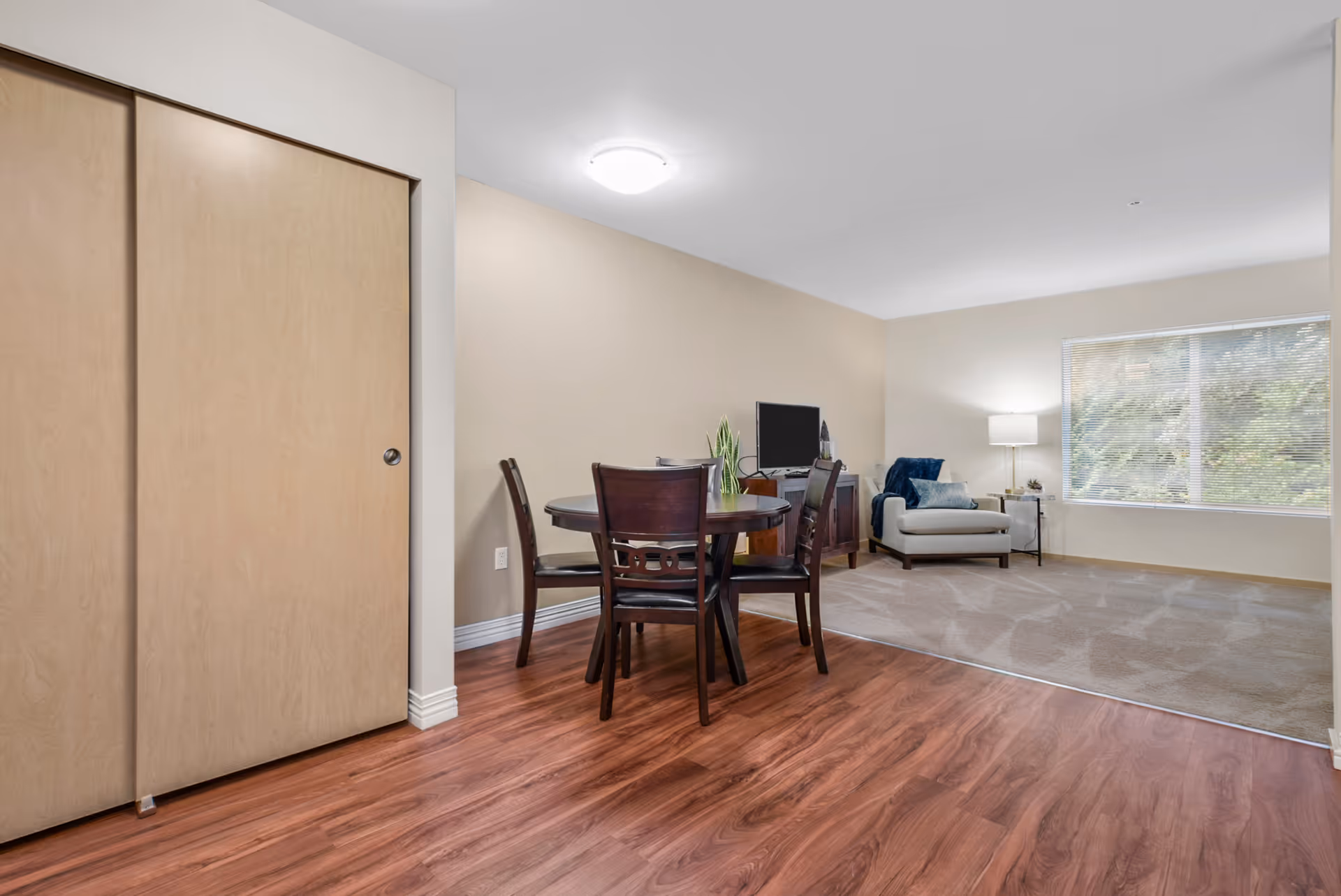 A bright and clean living area with a small round dining table and four chairs on wood flooring. Adjacent to the dining area is a carpeted lounge space with a single armchair, a side table with a lamp, and a flat-screen TV on a stand. A large window with blinds allows natural light to fill the room. There is a closed sliding door closet on the left side.