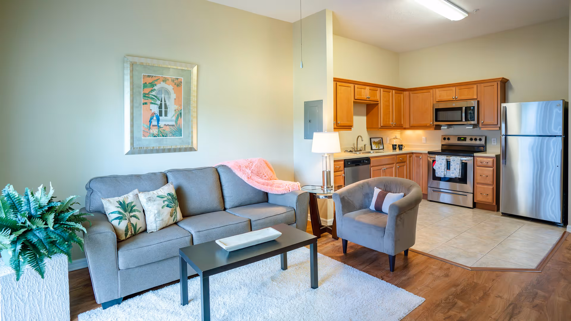A cozy living room area with a gray sofa adorned with tropical-themed pillows and a pink throw blanket. Next to the sofa is a small round glass side table with a lamp. A gray armchair with a decorative pillow faces a black coffee table on a white rug. The living room opens into a kitchen with wooden cabinets, stainless steel appliances including a refrigerator, microwave, stove, and dishwasher. A framed tropical artwork hangs on the wall above the sofa, and a large potted plant is placed on the left side of the sofa.