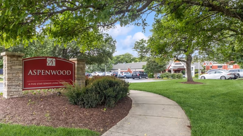 Outdoor view of Aspenwood Senior Living Community showing a red sign with the community name and slogan, a curved sidewalk, green grass, trees, parked cars, and buildings in the background under a partly cloudy sky.