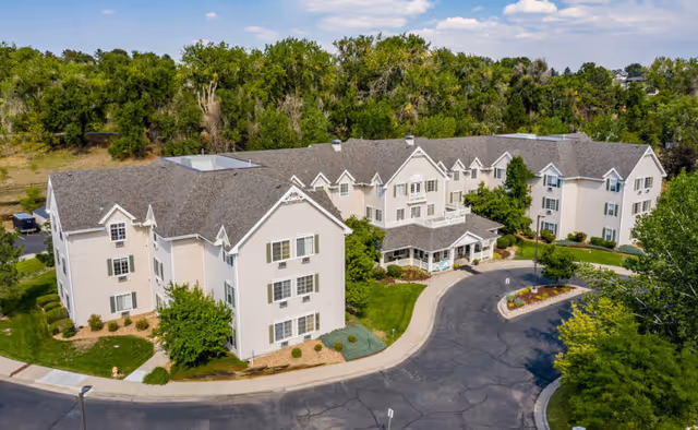 Aerial view of a large, multi-story senior living facility building with beige exterior walls and a gray roof, surrounded by greenery and trees. The building has multiple windows and a curved driveway leading to the main entrance. The sky is partly cloudy.