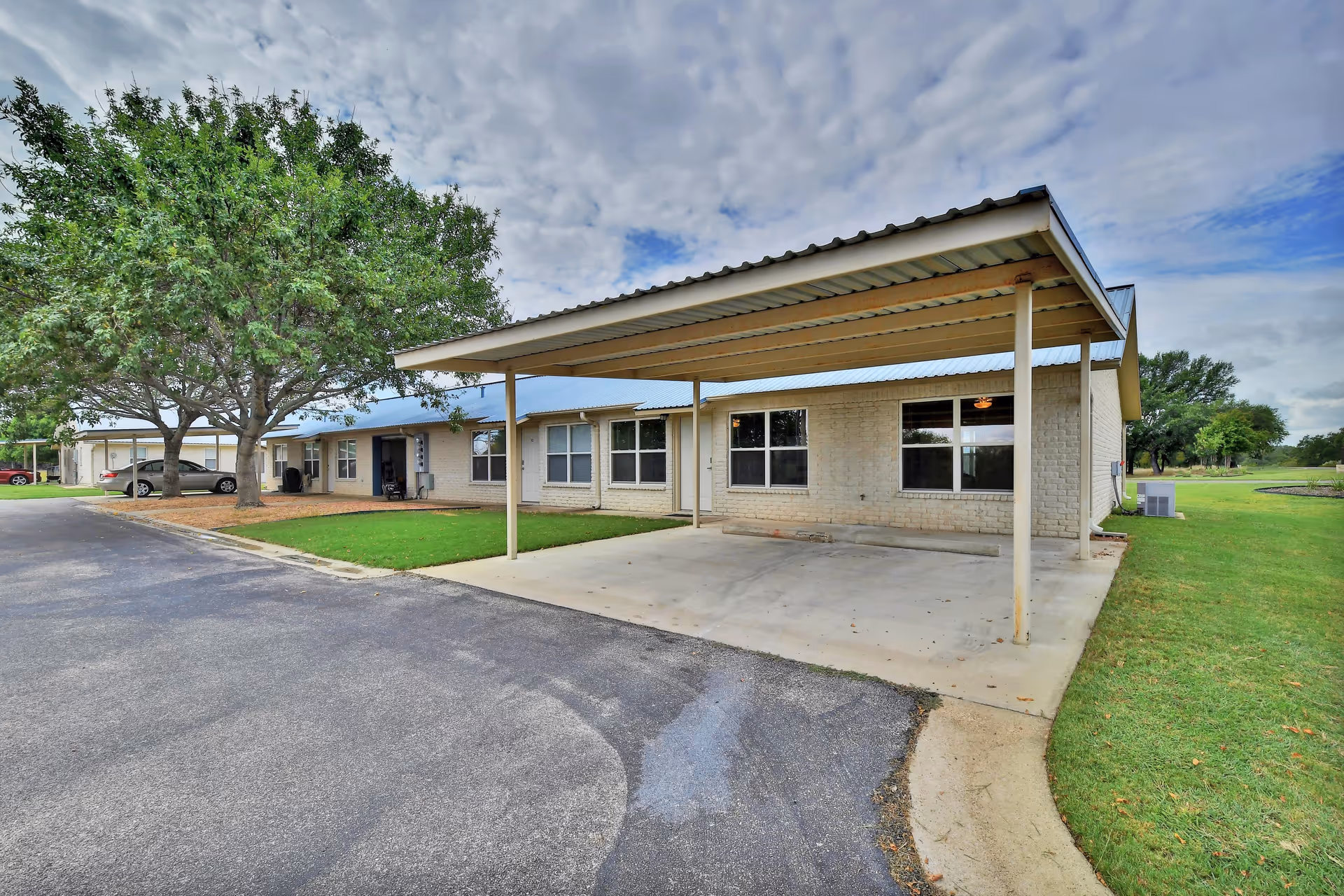 Exterior view of a single-story building with a covered carport, several windows, and a tree on the left side. The building is surrounded by a paved driveway and green grass under a partly cloudy sky.