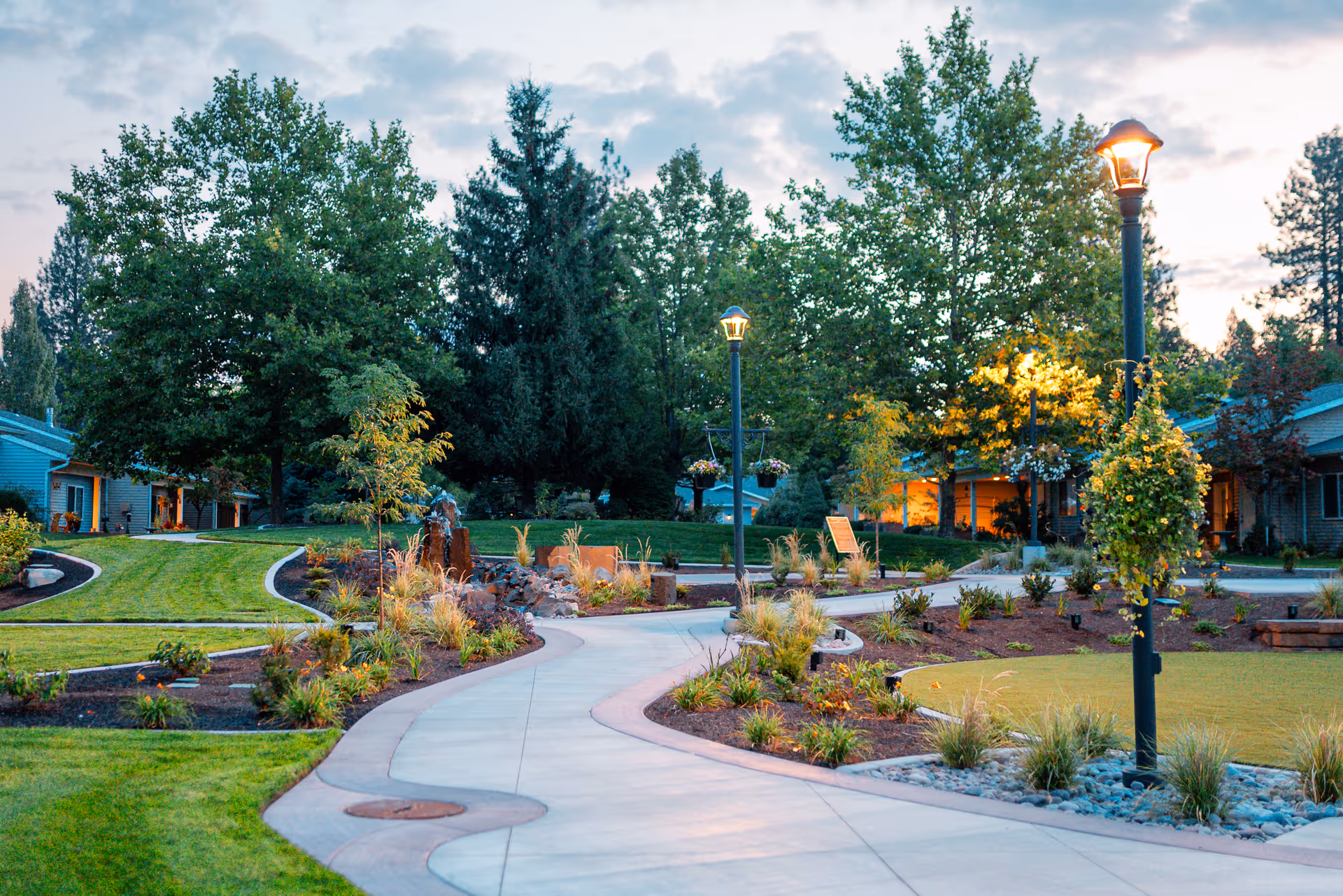 A winding concrete pathway through a landscaped garden area with green grass, various plants, and trees. There are lamp posts with hanging flower baskets lit up along the path. Residential buildings with warm lights glowing from inside are visible in the background during dusk.