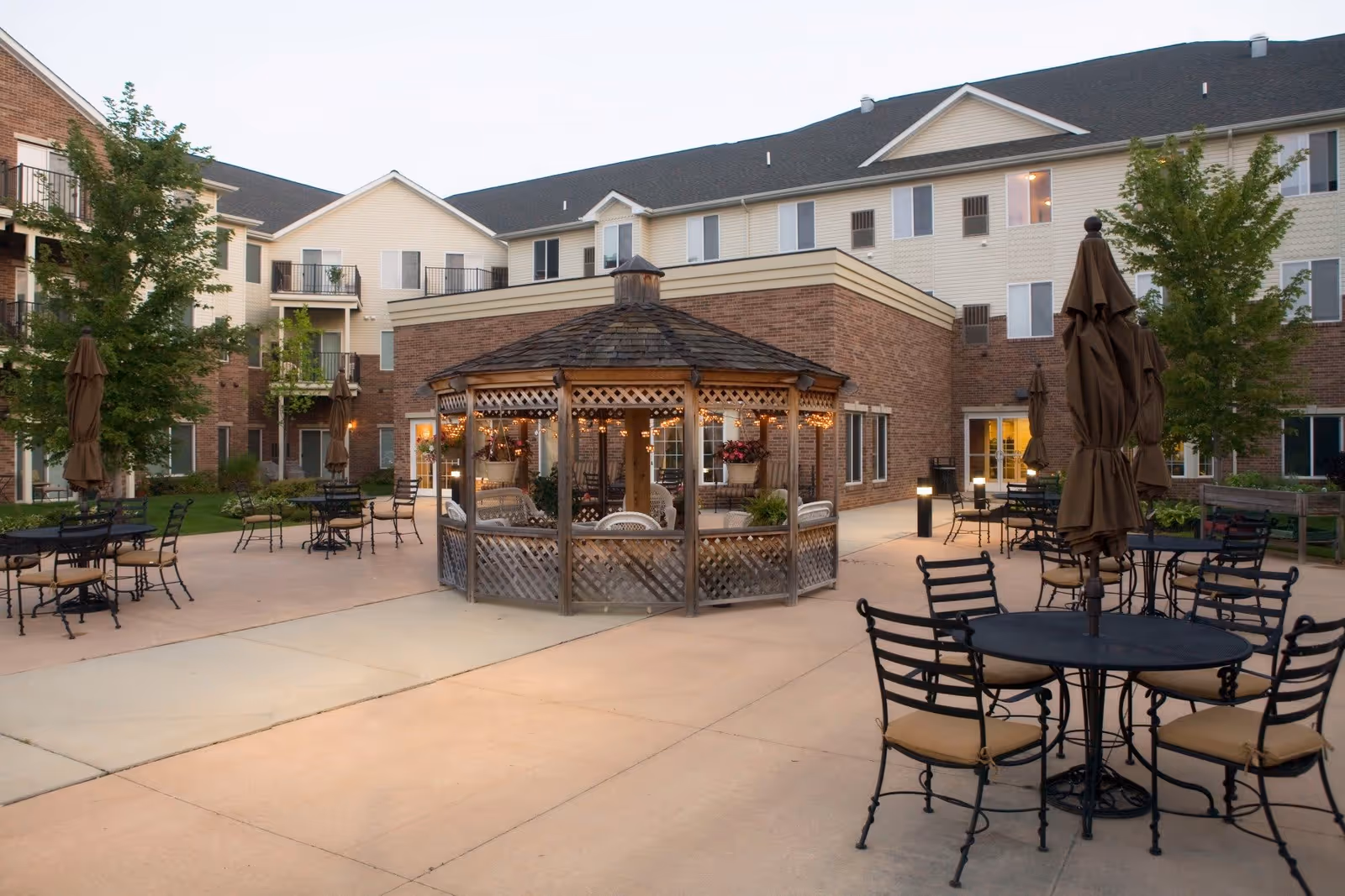 Outdoor patio area at American House Sterling Woods featuring a central wooden gazebo decorated with string lights, surrounded by multiple round tables with chairs and closed umbrellas. The background shows a multi-story building with balconies and windows, along with some trees and greenery.