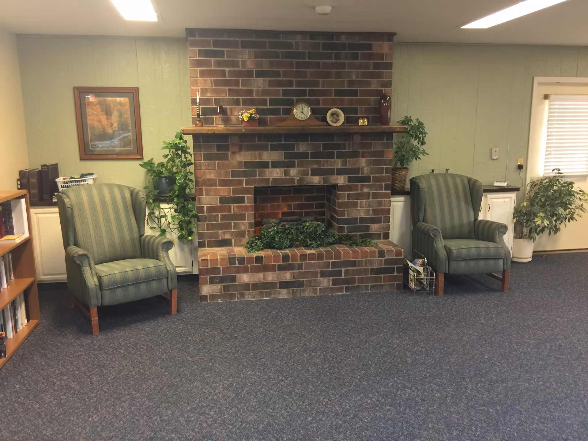 A cozy interior room featuring a brick fireplace with a wooden mantel decorated with a clock, small plants, and decorative items. On either side of the fireplace are two green striped armchairs. There are potted plants and bookshelves along the walls, with a window covered by blinds on the right side.