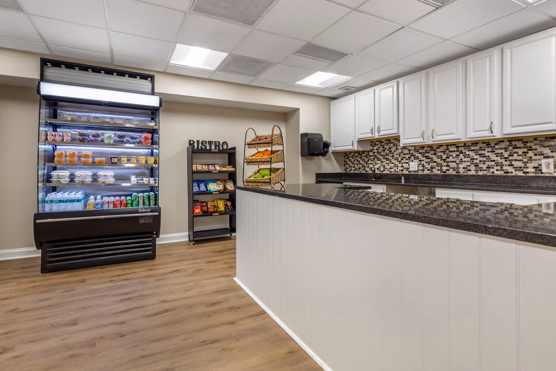 Interior view of a small bistro area in a retirement community with a refrigerated display case stocked with beverages and food items, a shelving unit with snacks, a countertop with a black granite surface, white cabinets, and a mosaic tile backsplash.