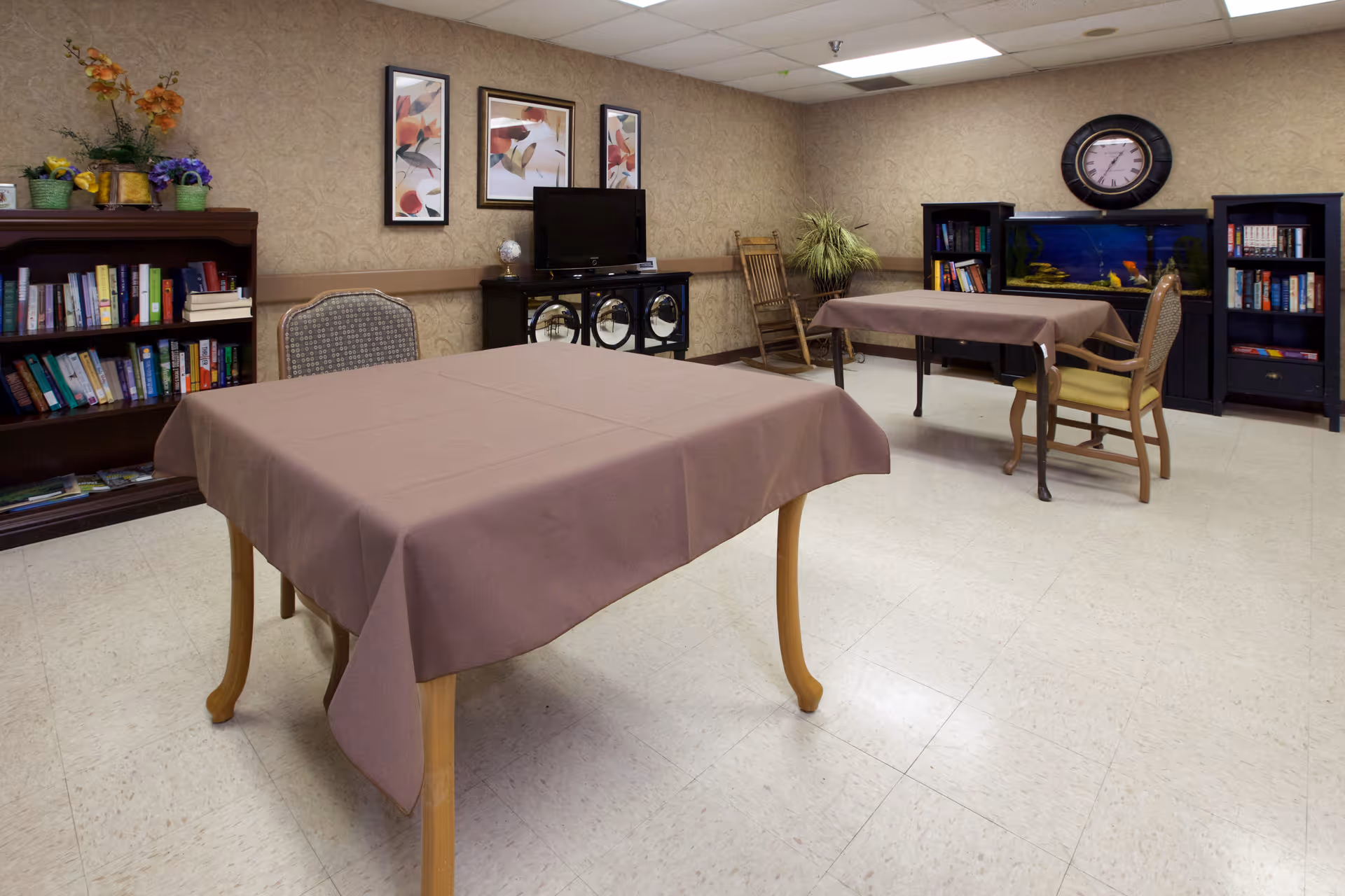 Community activity/dining room with two tables covered in brown tablecloths, bookshelves, a TV, fish tank and chairs.