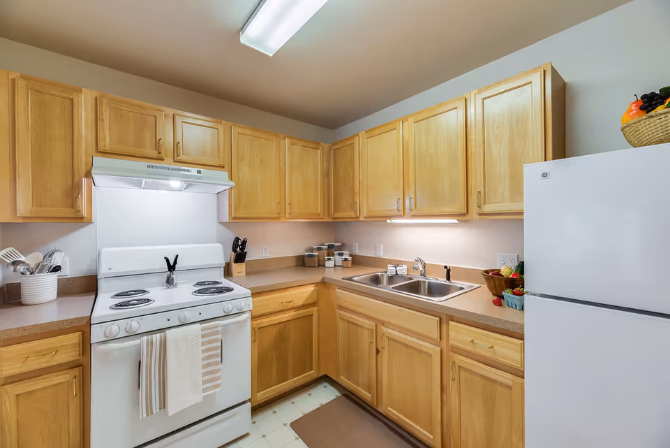 Well-lit kitchen with light wood cabinets, a white stove and refrigerator, double sink, and countertop accessories.