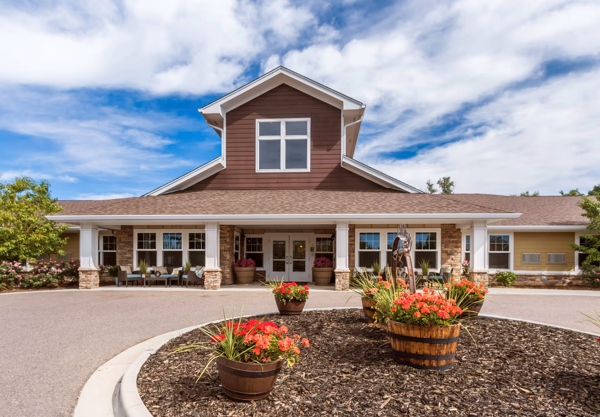 Front exterior view of a single-story building with a peaked roof and large windows. The entrance is covered by a porch supported by white columns with stone bases. In front of the building, there is a circular landscaped area with flower pots containing red flowers and green plants. The sky is partly cloudy with blue patches.