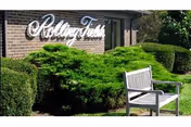 Outdoor view of the Rolling Fields Eldercare Community sign mounted on a brick wall, surrounded by green bushes and a wooden bench on a grassy area.