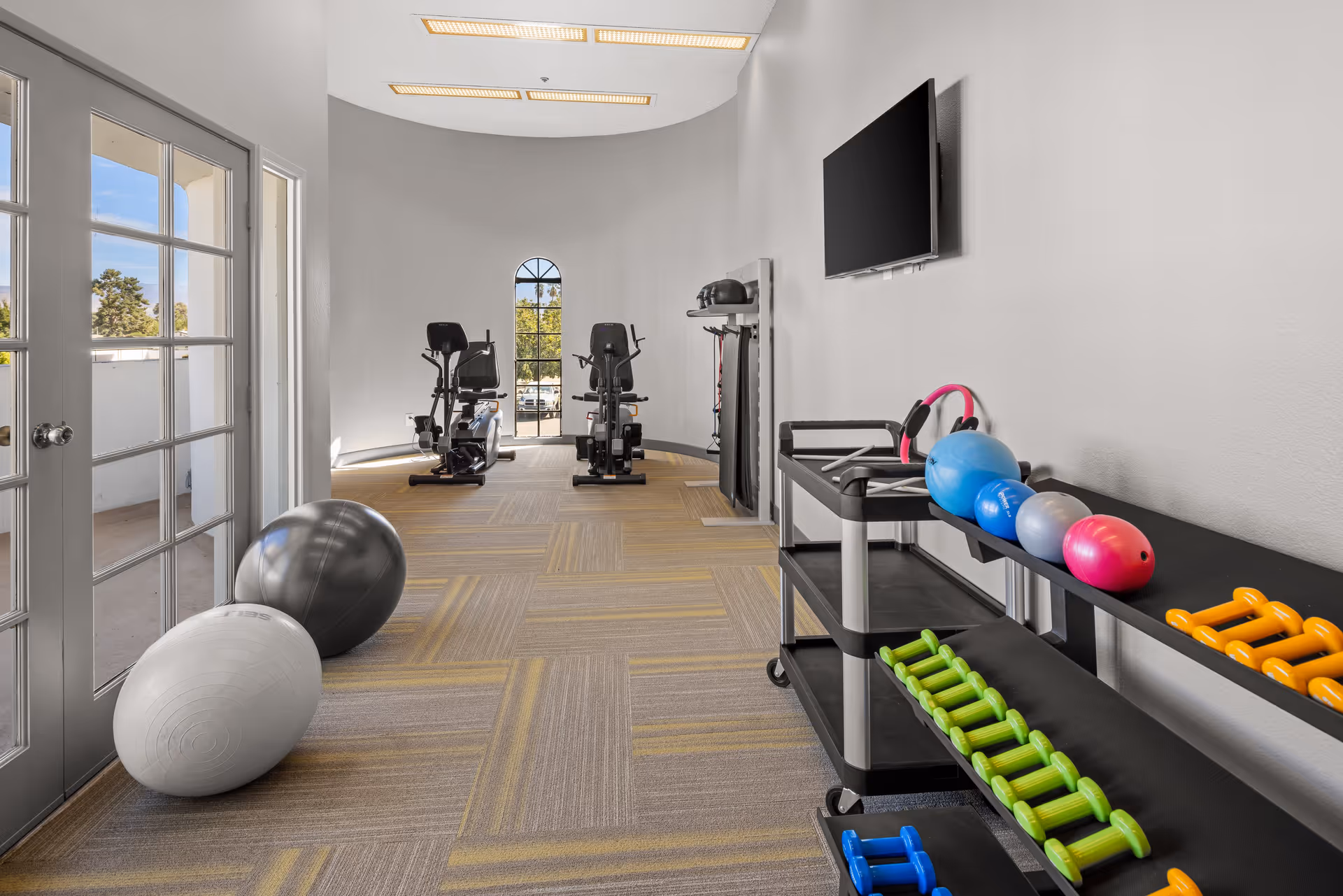 A fitness room with exercise equipment including two recumbent bikes, various dumbbells, kettlebells, and exercise balls. The room has large windows and glass doors letting in natural light, with a TV mounted on the wall.