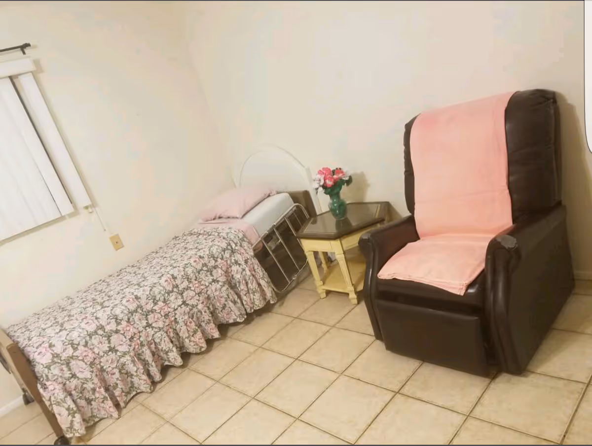 A small bedroom with a single bed covered in a floral bedspread and a pink pillow. Next to the bed is a small yellow nightstand with a glass top, holding a green vase with pink and white flowers. To the right of the nightstand is a dark brown recliner chair with a pink cover draped over the back and seat. The room has tiled flooring and a window with vertical blinds on the left wall.