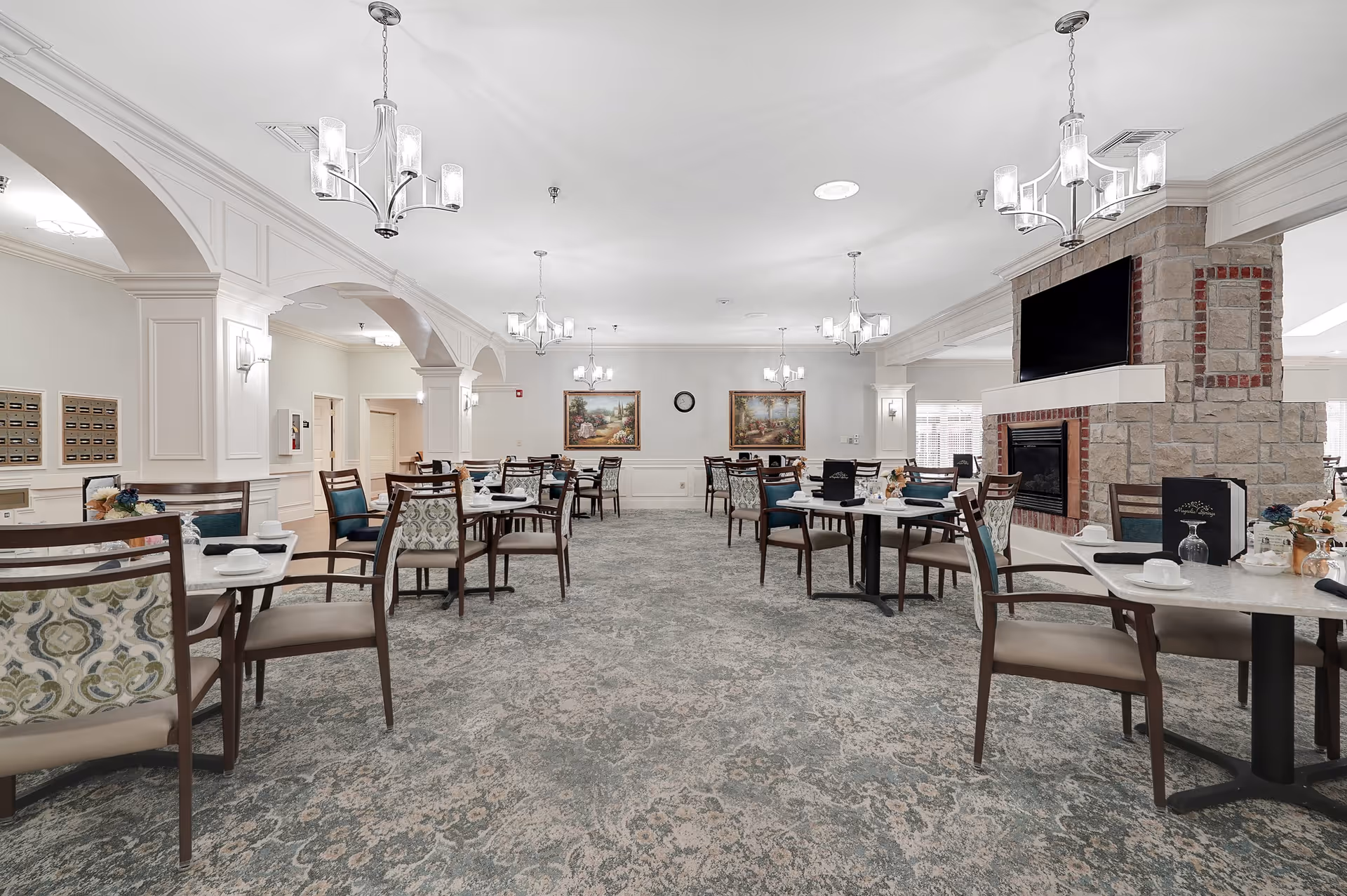 A spacious dining room with multiple tables and chairs arranged neatly. The room features elegant chandeliers hanging from the ceiling, a stone fireplace with a mounted TV above it, and framed paintings on the far wall. The carpet has a patterned design, and the walls are painted white with decorative molding.