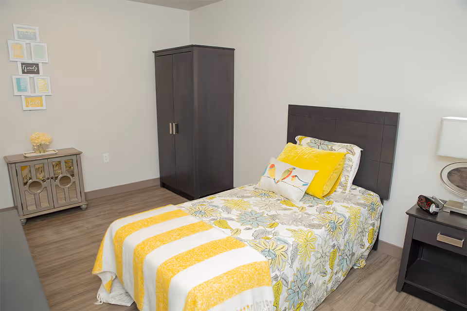 A neatly made single bed with floral and yellow bedding in a bright bedroom. The room features a dark wooden headboard, a matching dark wooden wardrobe, a nightstand with a lamp and clock, and a small cabinet with decorative items. The walls are light-colored with framed artwork hanging on one side.