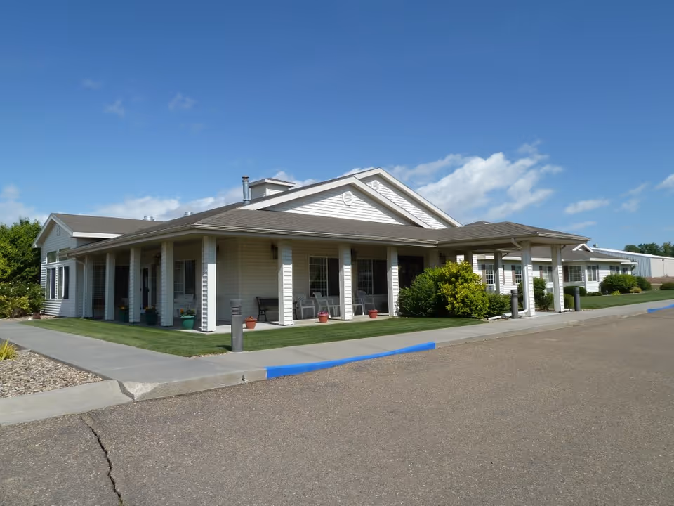 Single-story senior living facility building with a covered entry, columns, chairs, and landscaping under a blue sky.