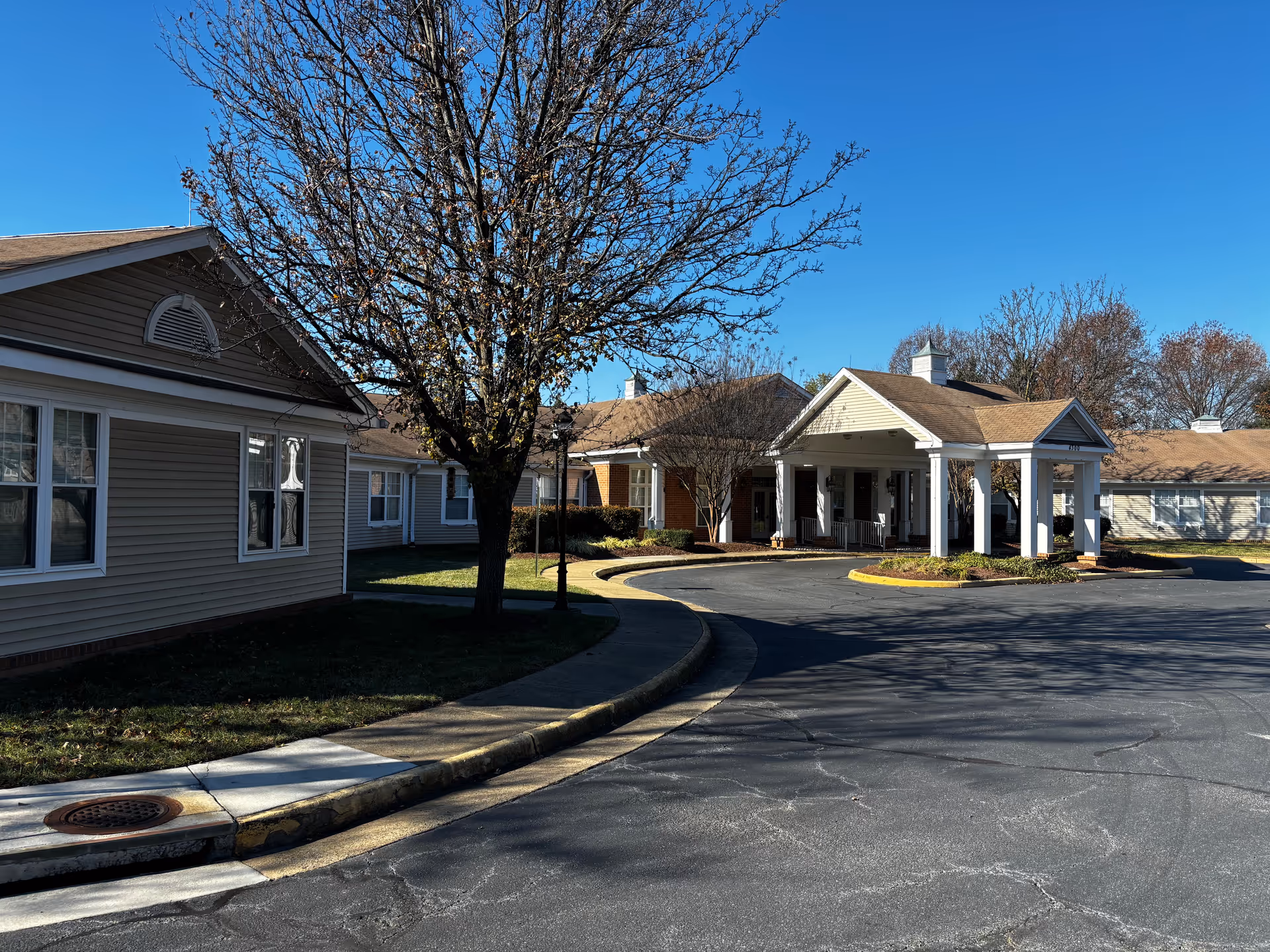 Exterior view of Sunrise of Fairfax senior living facility showing a driveway with a covered entrance, beige siding buildings, leafless trees, and a clear blue sky.