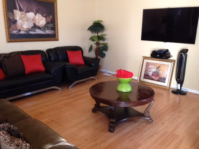 Living room with black leather sofas, a round wooden coffee table holding a green vase of red flowers, a wall-mounted TV, and framed artwork.