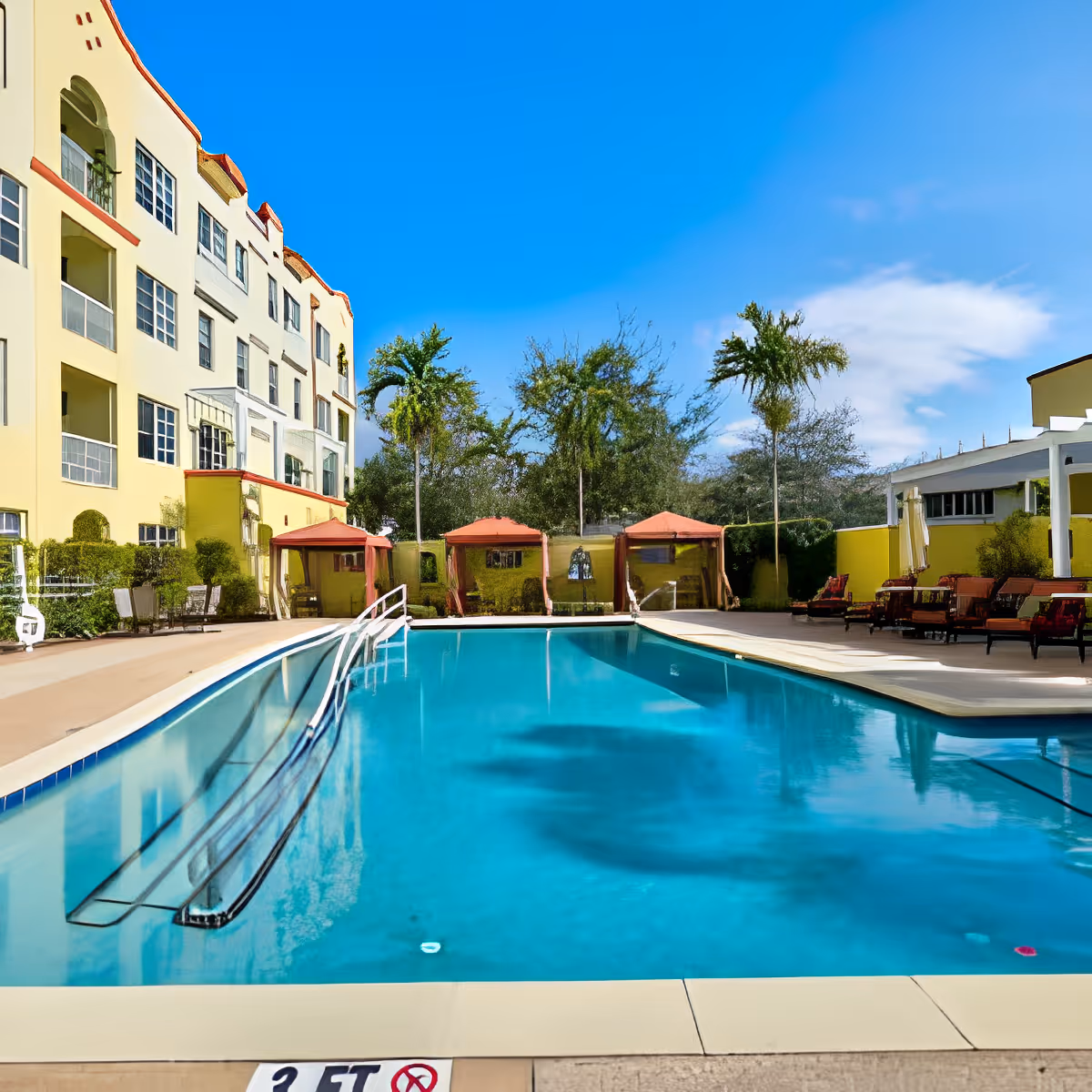 Outdoor swimming pool with lounge chairs, cabanas, and a multi-story yellow building under a blue sky.