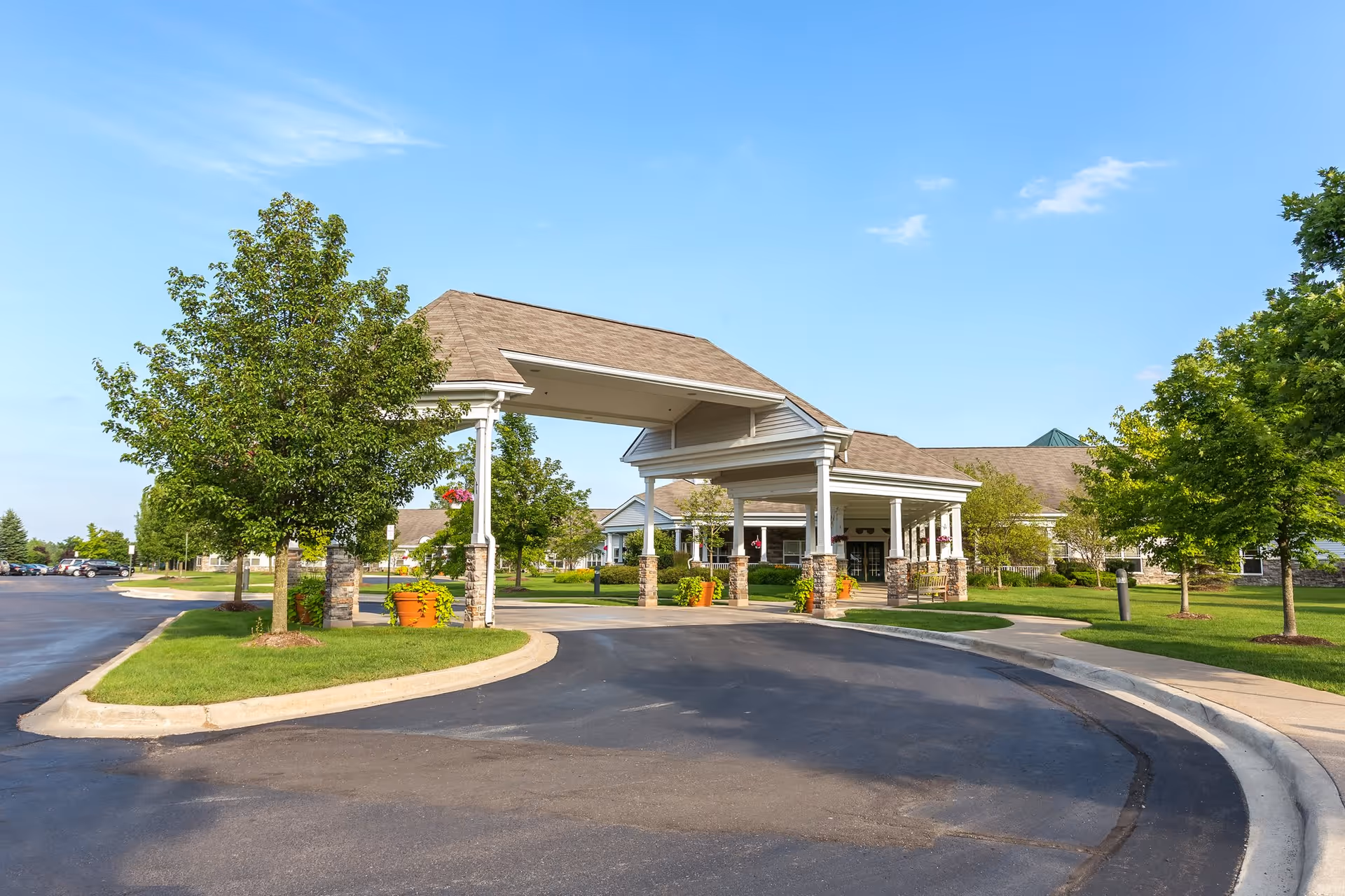 Entrance driveway with a covered porte-cochère at Independence Village of Oxford (Waterstone) facility, surrounded by green trees and well-maintained landscaping under a clear blue sky.