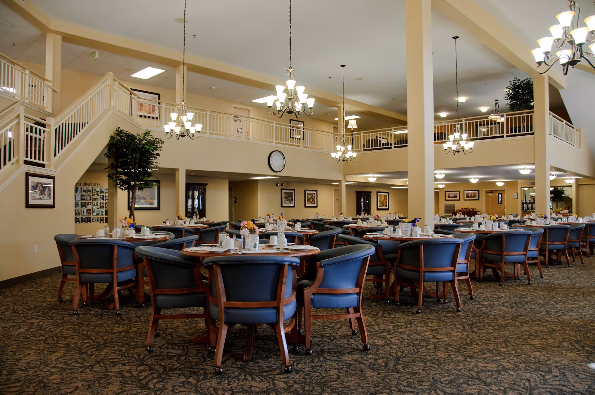 Spacious dining room in a senior living facility with multiple round tables set with cups, plates, and flower centerpieces. The room features blue cushioned chairs with wooden frames, chandeliers hanging from the ceiling, a carpeted floor, and a second-floor balcony with white railings overlooking the area.