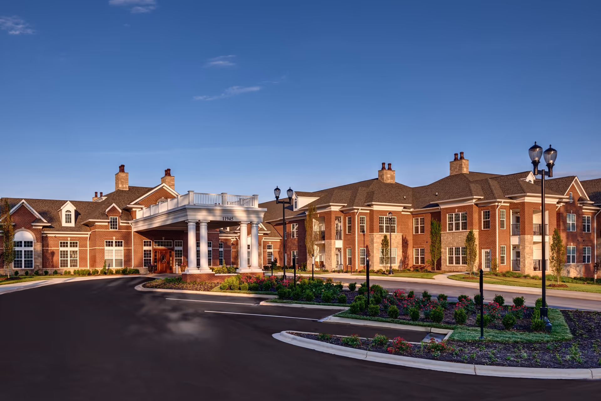 Exterior view of Waltonwood Providence, a large brick senior living facility with multiple chimneys, white columns at the entrance, landscaped gardens, and a curved driveway under a clear blue sky.