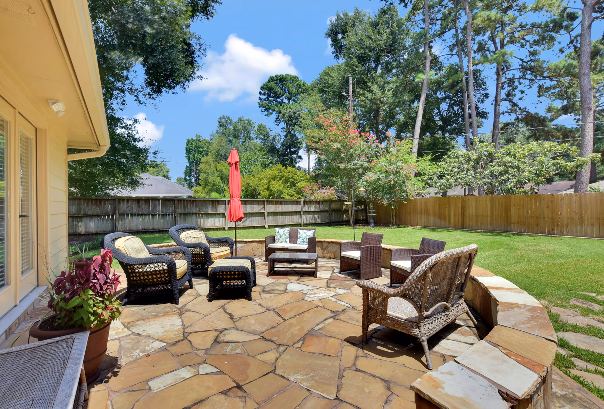 Outdoor patio area with stone flooring, surrounded by a wooden fence and green lawn. The patio features several wicker chairs with cushions, a small table, and a closed red umbrella. Trees and shrubs are visible in the background under a blue sky with some clouds.