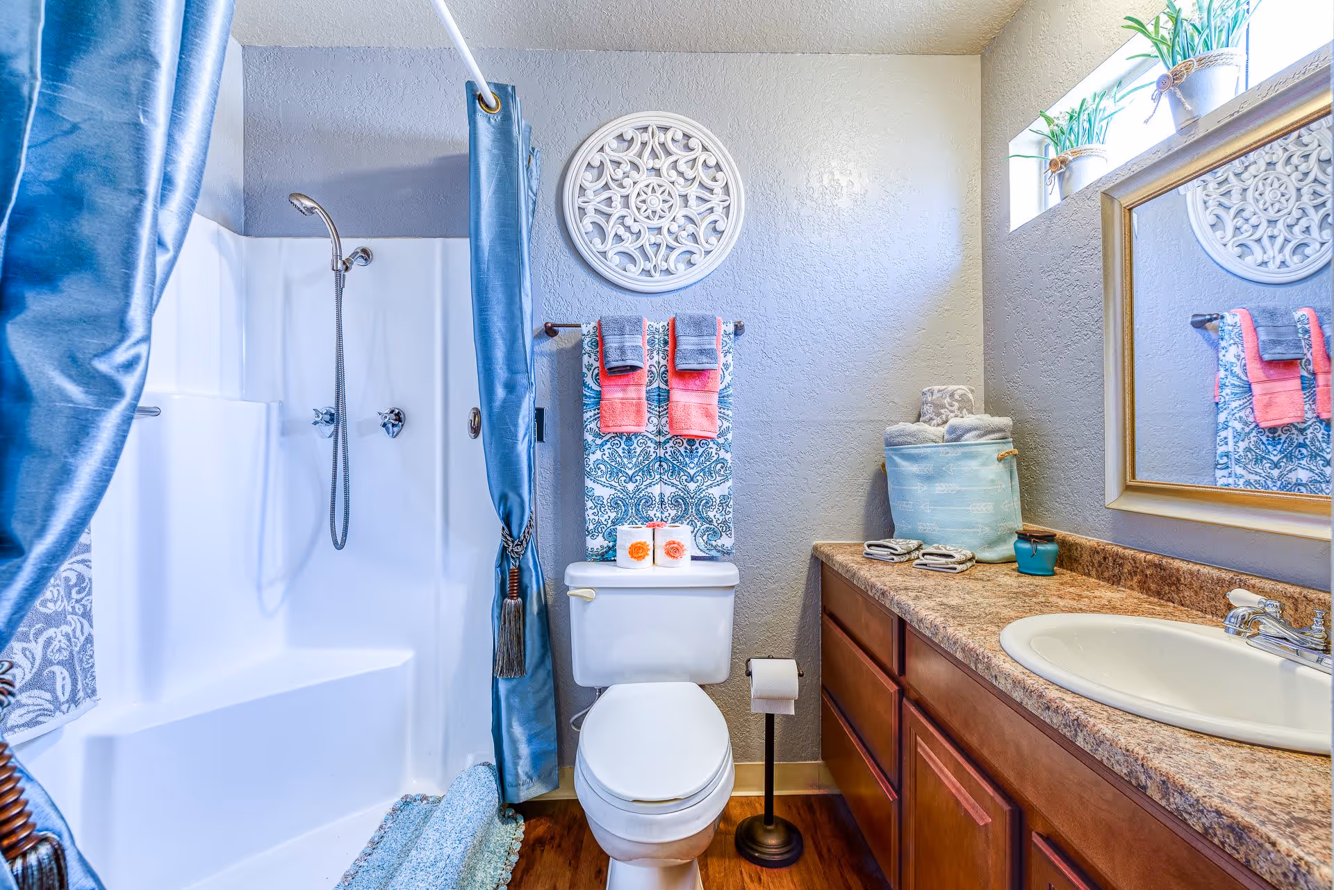 A bright and clean bathroom featuring a white toilet with decorative towels hanging above it, a shower area with a blue curtain, a countertop with a sink, and a mirror. The walls are light gray with a decorative round wall piece above the toilet. There are small potted plants on the windowsill providing natural light.