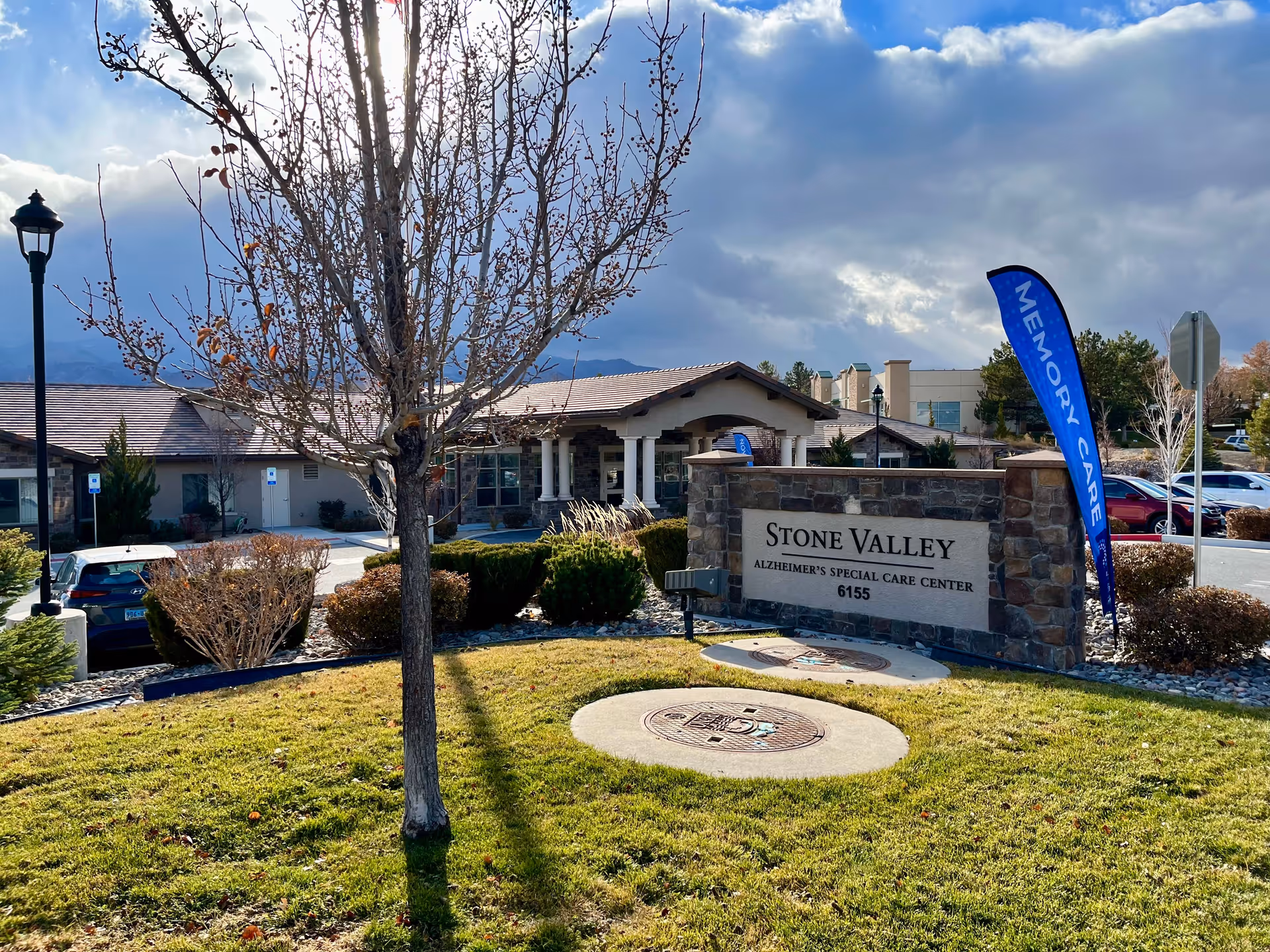 Exterior view of Stone Valley Alzheimer's Special Care Center with a stone sign displaying the facility name and address. A blue flag with the words 'Memory Care' is next to the sign. There is a tree in the foreground, bushes, parked cars, and a partly cloudy sky in the background.