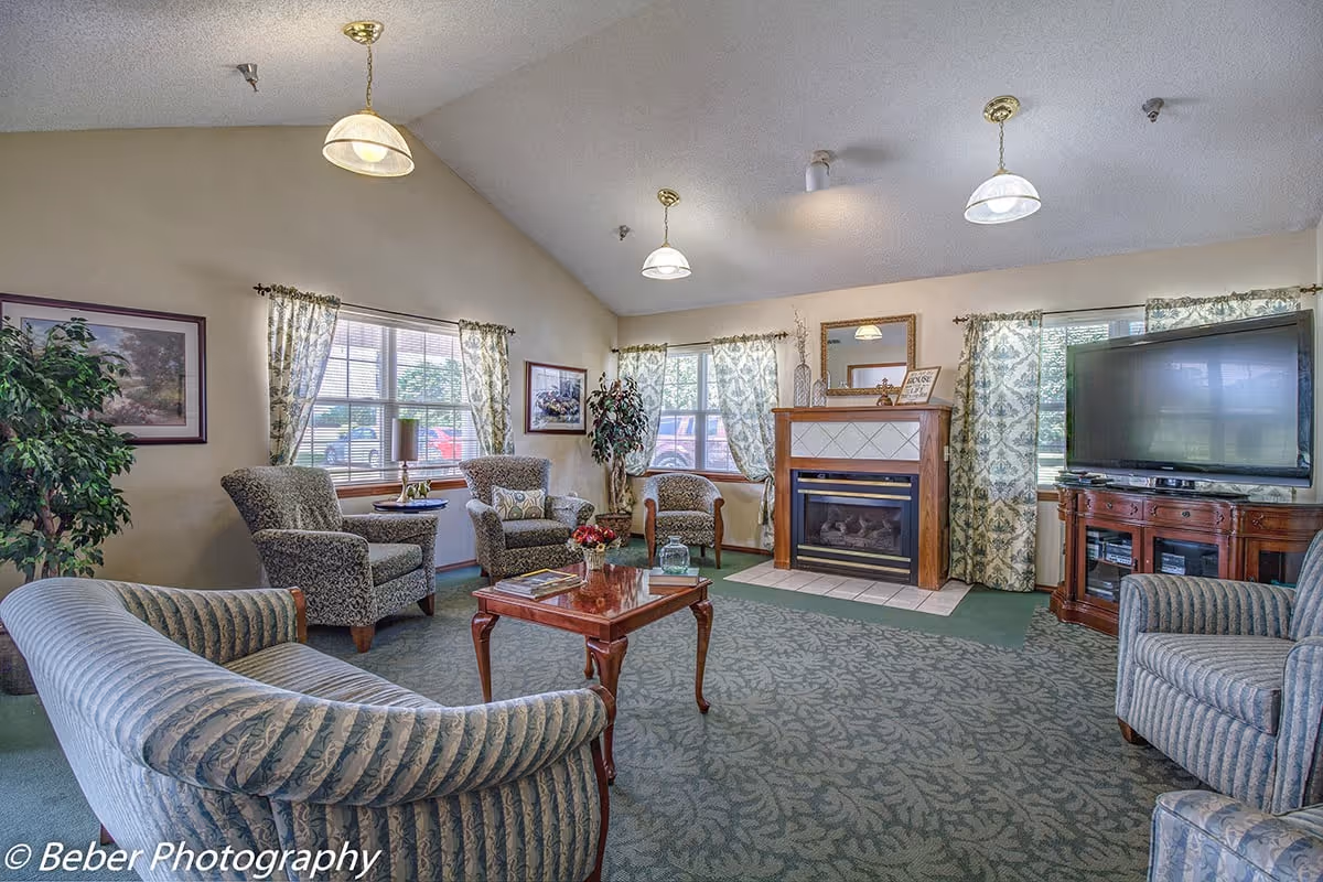 A cozy senior living facility common area with patterned armchairs and sofas arranged around a wooden coffee table. The room features a fireplace with a decorative mantle, a large flat-screen TV on a wooden stand, and windows with floral curtains letting in natural light. The carpet has a leafy pattern, and there are framed pictures and plants adding to the homey atmosphere.