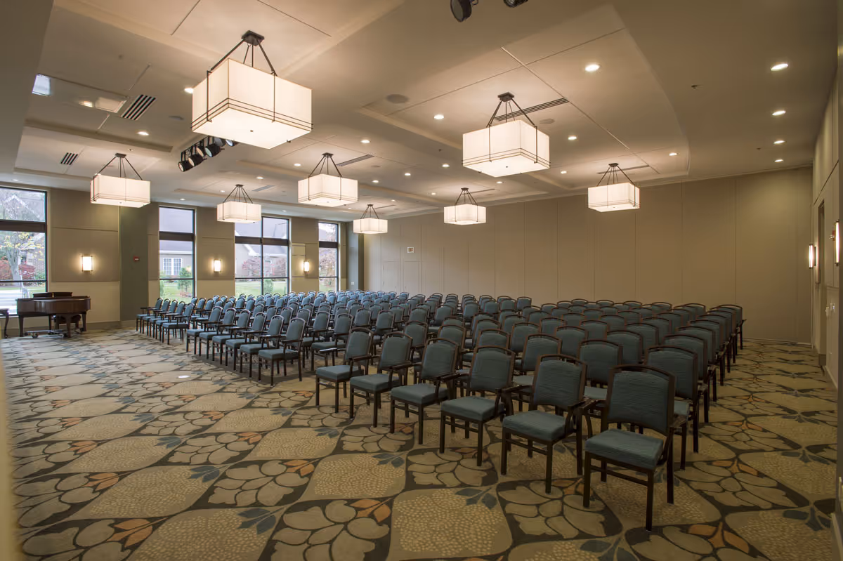 A large conference or meeting room with rows of blue cushioned chairs arranged facing forward. The room has large windows on one side letting in natural light, patterned carpet flooring, and multiple modern rectangular ceiling light fixtures. A grand piano is visible in the corner near the windows.