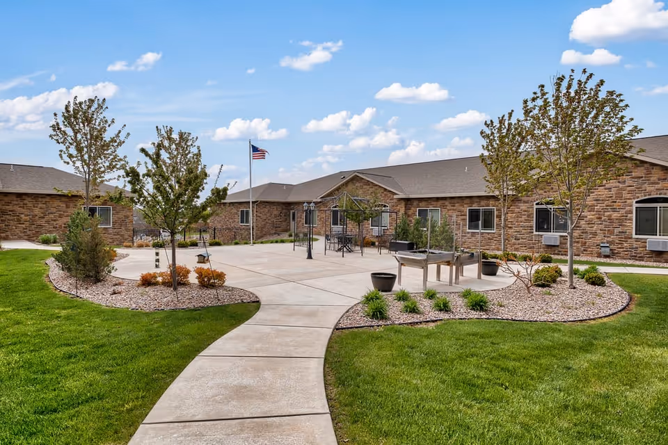 Courtyard with a paved walkway, landscaped beds, seating area and an American flag in front of low brick residential buildings under a blue sky.