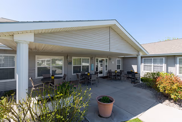Outdoor covered patio area at Avista Senior Living Twin Falls with several tables and chairs arranged for seating, surrounded by plants and shrubs under a clear blue sky.